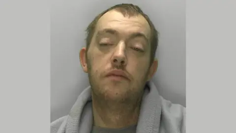 A police mugshot of James Smith. He is wearing a grey t-shirt with a grey blanket draped over his shoulders. He has short brown hair and a brown beard extending down the front of his neck. He is standing against a grey background and looking at the camera with his eyes half-shut and his head titled to the right.