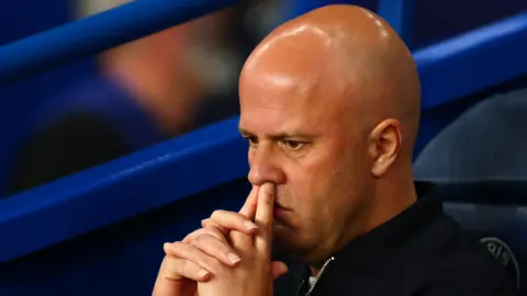 Headshot of Arne Slot, sitting on the bench, fingers on top lip, looking pensive