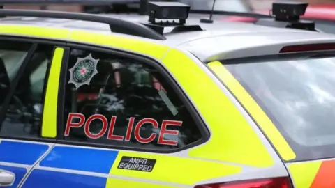 The back of a Police Service of Northern Ireland (PSIN) car. 
it is bright blue and yellow with the words: 'Police' and the PSNI crest on the window. Two cameras are at the top of the vehicle and a black sticker with the words: 'ANPR EQUIPPED' on the side of the car. 
