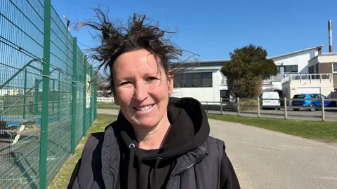 Vicky Barratt está de pie al aire libre en un día soleado, sonriendo a la cámara. Ella tiene cabello marrón oscuro azotado y lleva una sudadera con capucha negra en capas debajo de un chaleco de color oscuro. En el fondo hay una cerca de metal verde, un camino pavimentado, un moderno edificio blanco con grandes ventanas y varios autos estacionados. 