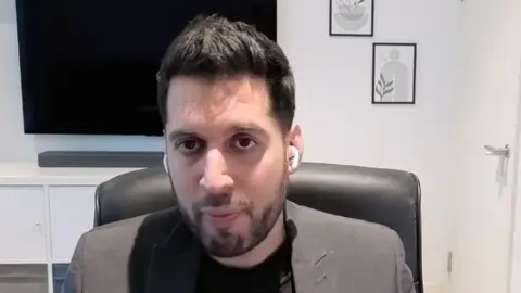 Rahan Arif, a man in a grey suit with a black T-shirt sits in a chair looking at the camera at his desk. He has white headphones in his ears.