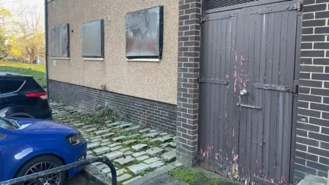 The exterior of the flat of the BBC ground floor where the closes were abused. The three windows have a brown metal mesh. The entrance is a brown double wooden door whose color is off and it is closed. In the pictures, the bumper of two park cars and the back part are also visible.