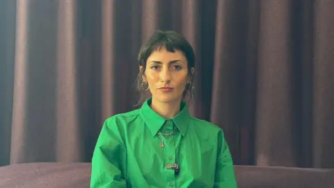 A woman with dark hair is sitting on a brown sofa in front of a brown curtain. She is wearing a green buttoned up shirt and is smiling at the camera