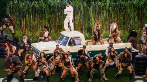 PA Media Musician Bad Bunny performing at the Super Bowl in the US. He is wearing all white and standing on a cream coloured truck while a large group of people are dancing around him.