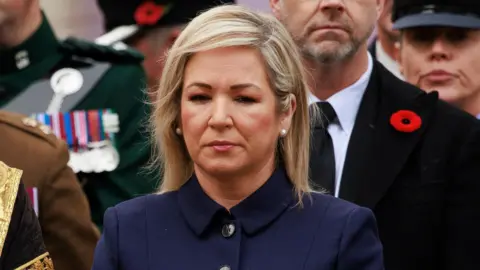 PA Michelle O'Neill looking solemn in navy suit jacket at remembrance event, men pictured in background are wearing dark clothes, war medals and a poppy