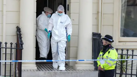 Forensic officers in white protective suits leave a cordoned-off building in Brighton as a uniformed police officer stands nearby.