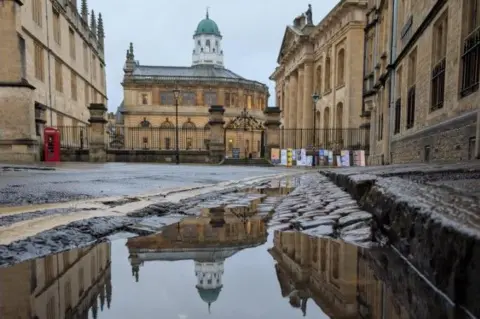 Lucie Johnson The cobbled kerb is lined with puddles. The Sheldonian Theatre and a red postbox are in the background.