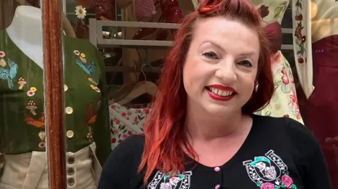 Owen Sennitt/BBC Jenny Bee Lee, a woman who is standing outside her fashion boutique Lady Bee Loves which is located in the Royal Arcade in Norwich. She has red hair and is wearing a black cardigan and red lipstick.