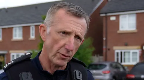 A photo of a man looking seriously to the side of the camera. He is an immigration enforcement officer. He has grey hair and a black vest on. 