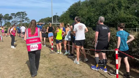Alice Harvey People line up at the end of the Jersey Parkrun. Volunteers wearing pink high-vis jackets are stood on the opposite side of a red and white chain fence where the runners are lined up.