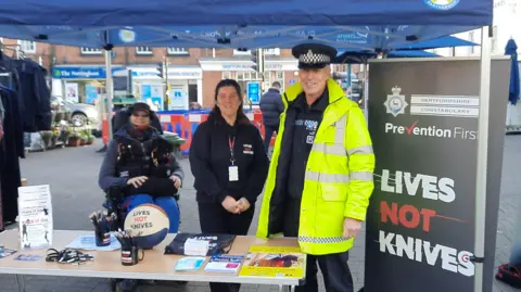 Hertfordshire Constabulary A stall run by Hertfordshire Constabulary at St Albans market. There three people looking straight at the camera, one officer, in full uniform with a high vis jacket. They have a table with pens, lanyards and leaflets on it. You can see market stalls behind and banner to the right. They are under a blue gazebo. 