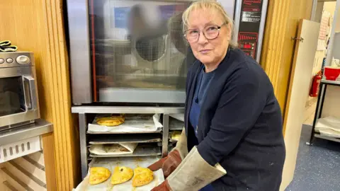 Susan has blonde hair and is wearing glasses. She is holding a tray of pasties.