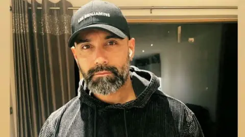 Met Police Mehmet Koray Alpergin stands in front of a window with dark gold curtains pulled to the side. He is wearing a black baseball hat, in-ear headphones and has a black jumper on.