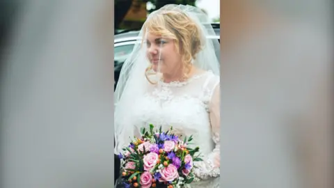 Charmain Speirs is wearing a white wedding dress with a veil over her head. She's stood in front of a car and is holding a bouquet of pink, purple, and orange flowers.