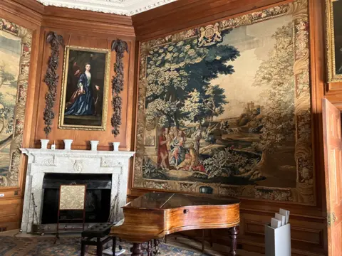 BBC/Rachel Royce An ornate historic room featuring large wall tapestries depicting detailed landscape scenes with trees and figures. A polished wooden grand piano stands in the centre foreground on an intricately patterned rug. Against one wall is a carved white fireplace with decorative urns on the mantel and a framed portrait above it. The wood‑panelled walls, high ceiling with plaster detailing and classical furniture create a richly decorated period interior.