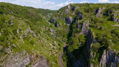 Cars parked along a winding road running through Cheddar Gorge. The road is quite narrow and there is someone walking on the pavement on the right hand side of the road.