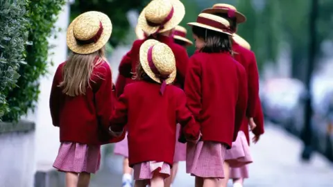 A group of schoolgirls are walking holding hands with their back to the camera. They are wearing red blazers, red gingham dresses and straw hats with a red bow