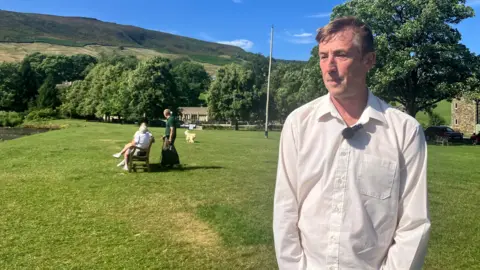 Richard Edwards/BBC A man standing on a village green, on the edge of a river. The hills are in the far distance and a dog is walking towards the camera. The man has brown hair and is wearing a white shirt.