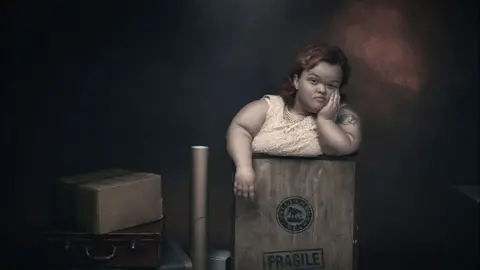 Debbie Todd A woman with dwarfism has a bored expression on her face as she stands in a box marked fragile