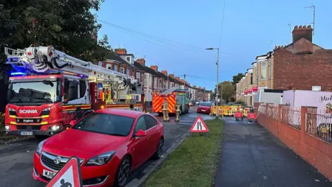 Two fire engines on a road, with terraced housing either side. A property on the road has no roof and smouldering embers can be seen. 