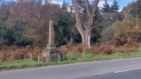 Carmelo Garcia A view of a road - woodland behind it and a stone obelisk monument on the verge.