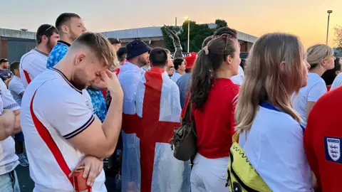 A man wearing an England football top has his head in his hands. He is surrounded by a large group of football fans who appear to be looking at a large screen.