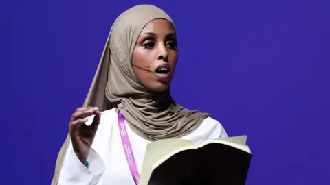 A young woman with a light brown headscarf and a white outfit is speaking with a small wire microphone near her mouth. She is gesturing with one hand and in the other is holding a black book
