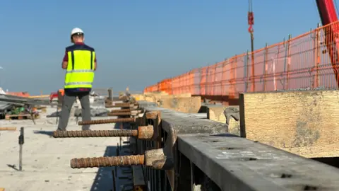 BBC Un trabajador de la construcción con un chaleco amarillo neón y un casco blanco se encuentra junto a un concreto del que sobresalen tornillos mientras trabaja en el nuevo proyecto ferroviario en Lituania.