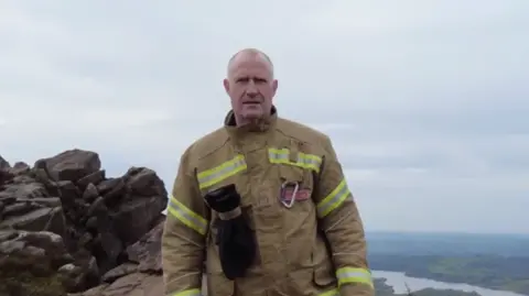 Staffordshire Fire and Rescue Service A man with a bald head in a brown firefighter over-suit standing on rocks over a valley below