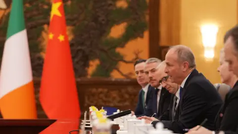 PA Media A picture of Micheál Martin, a man with thin grey hair, wearing a suit and sitting at a long table with other people in formal attire. An Irish and Chinese flag are in the background.