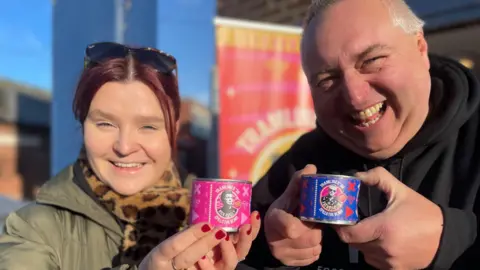 A woman with red hair wearing a thick green winter coat stands next to a jolly-looking man in a black hoodie. They are both holding up miniature cans of beans featuring pictures of Rick Astley and Fatboy Slim.