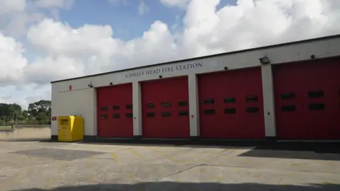 The outside of the current Camel Head Fire Station in Plymouth. The stations name is written on a sign above four large red doors. 