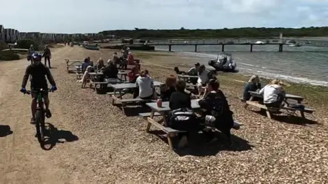 Google Street View of picnic benches on a beach on Mudeford sandbank. It is a sunny day and the benches are filled with people. Two cyclists are passing on the path between the beach and a long row of beach huts.
