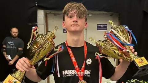 Family handout Alex Eastwood is holding three gold cups and has a medal around his neck. He is wearing a black T-shirt and is starring directly at the camera.