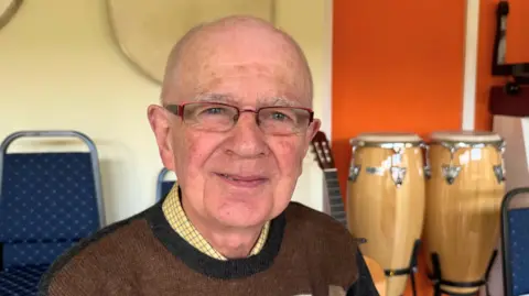 BBC/Emily Johnson An older man wearing glasses and a brown sweater sits in a room with drums, a guitar and rows of blue chairs behind him.