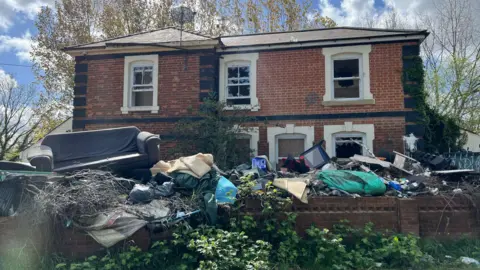 Thomas Magill/BBC The old station, now with broken and boarded up windows. All along the front of it, is flytipping - mattresses, white goods, blankets, and all kinds of rubbish. 
