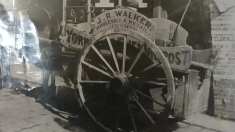 Suki Walker A black and white image, taken in the late 19th Century, showing a horse and cart. The cart has a sign on it stating: J.R.Walker. Wholesale and Retail. Newsagent'. There is also a sign stating: Yorkshire Evening Post 