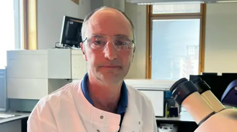 John Fairhall/BBC Dr Nick Tucker wearing a white lab coat in front of the twin eyepiece lenses