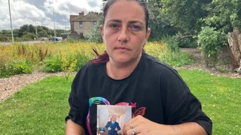 Stephanie Boyce-Williams is wearing a black t-shirt and has dark hair pulled back in a ponytail. She is sat in a garden area and is looking at the camera holding a photograph of Alan.