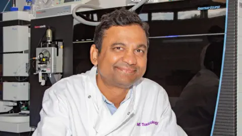 MRC Toxicology Unit Dr Patil is wearing a white lab coat with MRC Toxicology Unit printed on the left breast pocket in purple. He is sat in a lab with equipment behind him. He is smiling and has short dark brown hair and brown eyes.