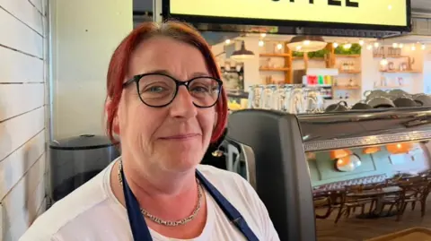 BBC Women with red hair and wearing black circular glasses. A sign that says coffee is behind her, along with a coffee machine.