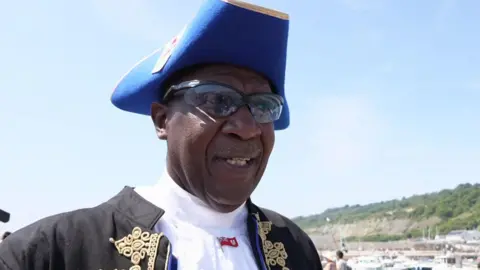 Ed Christopher is wearing traditional town crier uniform in black and blue, along with sunglasses.