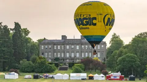 Claire Hartley A solitary hot-air balloon hovers just above a number of tents in the grounds of of a country estate. The main building can be seen in the background.