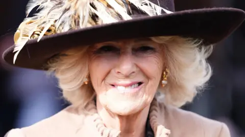 PA Media Queen Camilla on day two of the 2026 Cheltenham Festival at Cheltenham Racecourse. She is wearing a beige coat and a dark hat with feathers on.