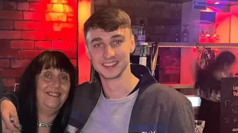 A woman with black hair and a fringe smiles at the camera is stood next to a teenager who has dark hair and is wearing a dark grey jacket and a light grey jacket