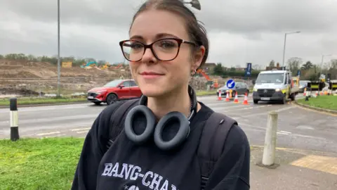 Henry Godfrey-Evans/BBC A woman with thick glasses and headphones round her neck smiling with a few cars waiting by roadworks