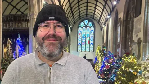 Alexander is wearing a hat and glasses and standing in front of an array of colourful, sparkly trees