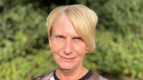 Laura Foster/BBC A head and shoulders image of a woman. She is standing in front of a green bush and looking into the camera.