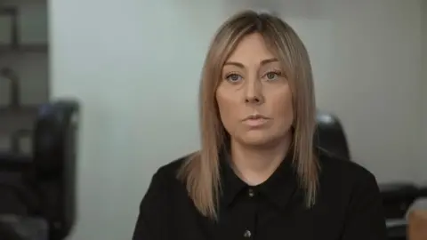A woman with dark blonde, shoulder-length hair. She is wearing a black button-up collared blouse and has a serious expression on her face.