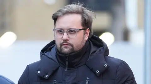 PC Liam Newman on his way to court wearing a black, hooded winter coat that is zipped up to to the chin with the hood down. He has straight fair hair worn in a side parting and is sporting a short beard. he is wearing clear rimmed spectacles with black and he is looking ahead, his gaze to the right of the camera